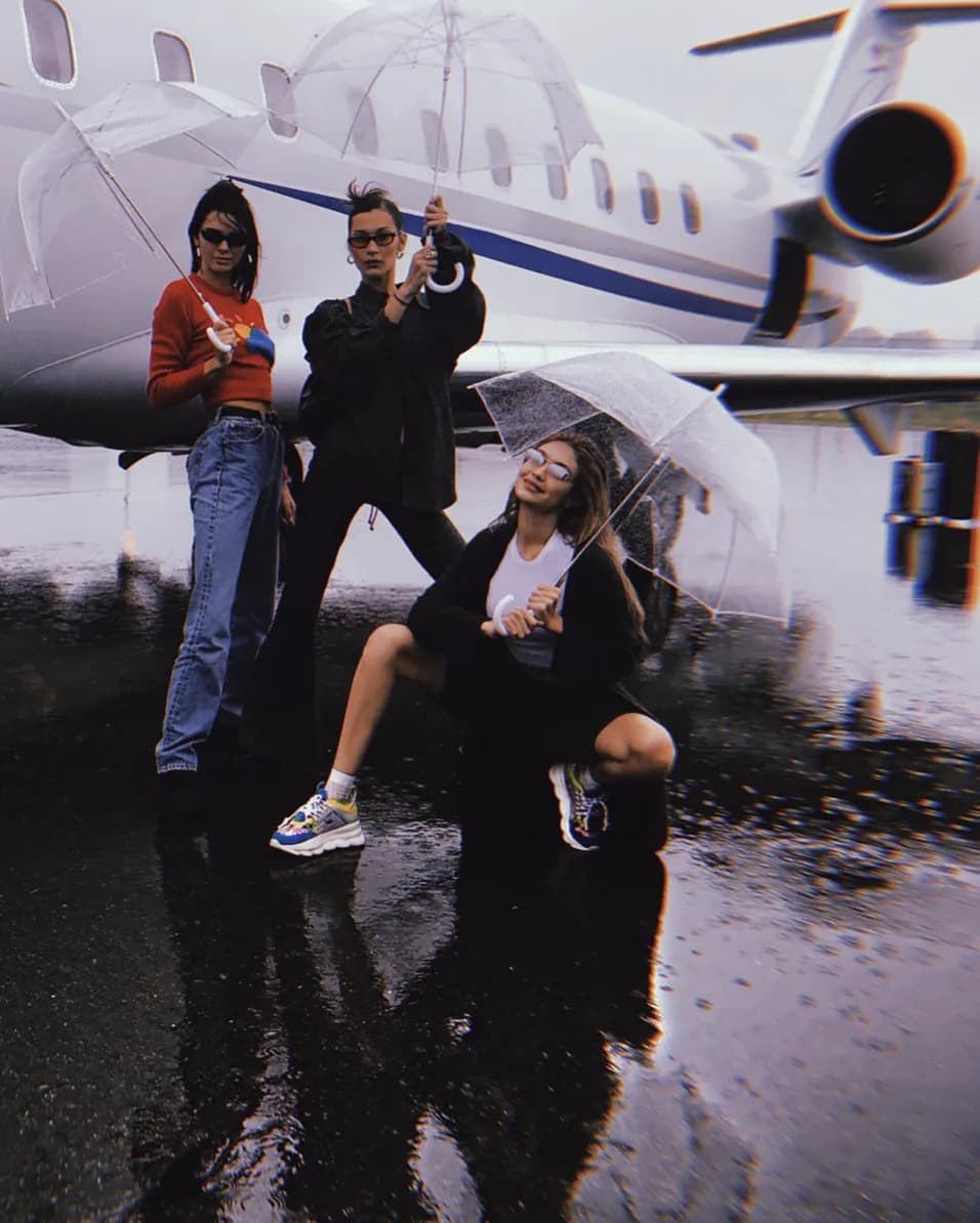 Bella Hadid, Gigi Hadid and Kendall Jenner under clear umbrellas on rain-slicked tarmac beside a private jet, shot for PRVT